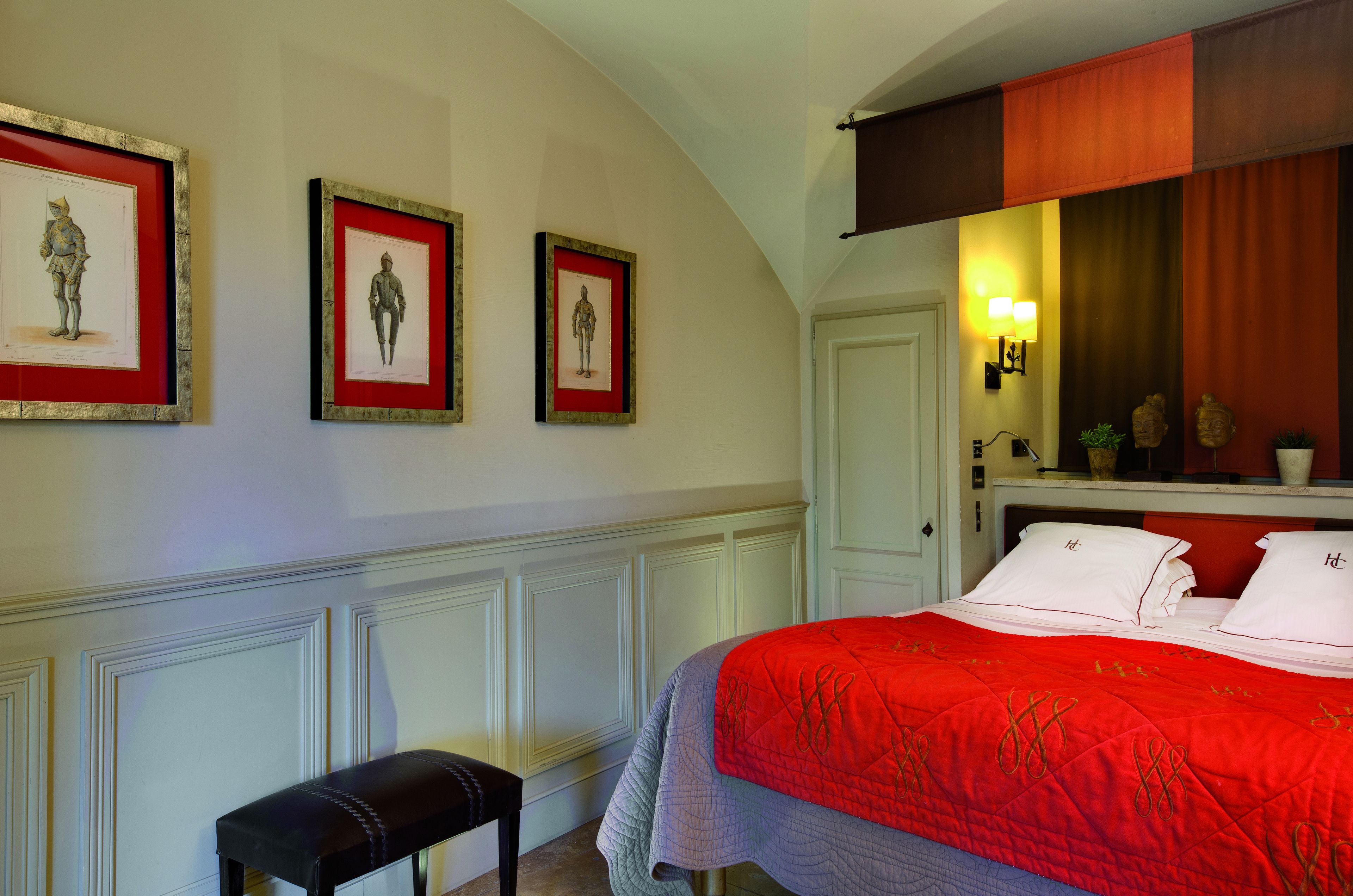 A bedroom with a red bedspread, a bench, and three framed pictures on the wall.