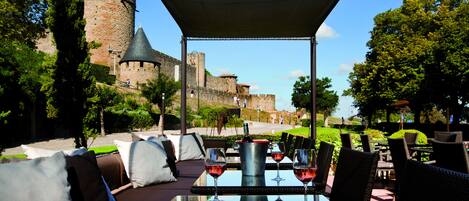 Un espacio para comer al aire libre con vistas a un castillo histórico, amueblado con mesas y sillas, y una botella de vino y copas sobre la mesa.