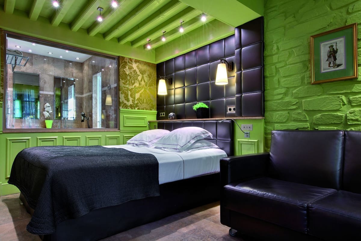 A room with a bed, a black leather sofa, a green accent wall, and a large mirror.