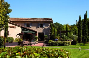 Um hotel com telhado de azulejos, entrada coberta e um jardim bem cuidado, com sebes e árvores.