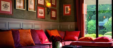 A room with a sofa set, framed artwork on the wall, and a view of greenery outside.