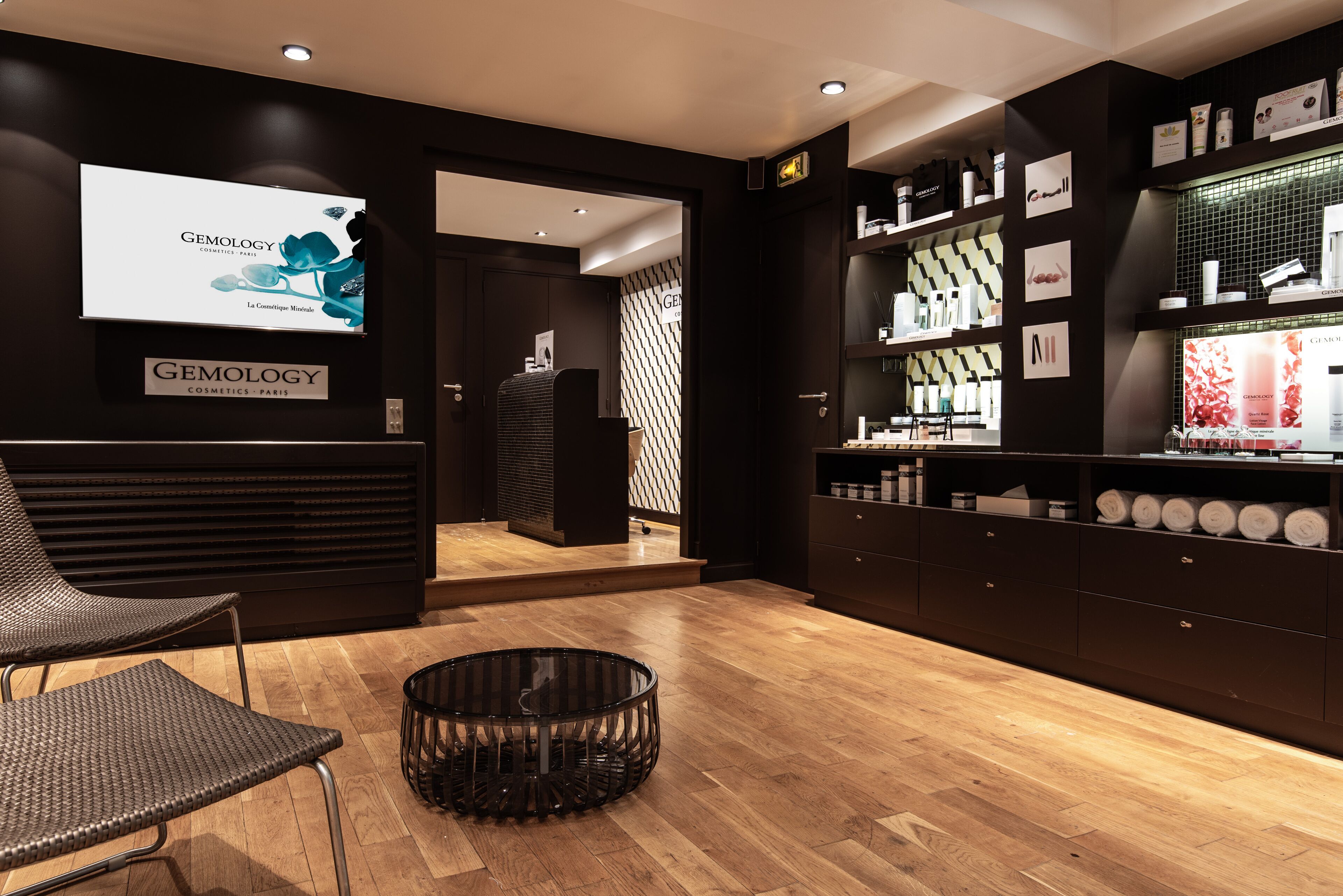 Un interior moderno con una paleta de colores oscuros, que incluye una zona de recepción con el letrero de 'Gemology Cosmetics Paris', un área de descanso con una silla de mimbre y una exhibición de productos en estanterías.