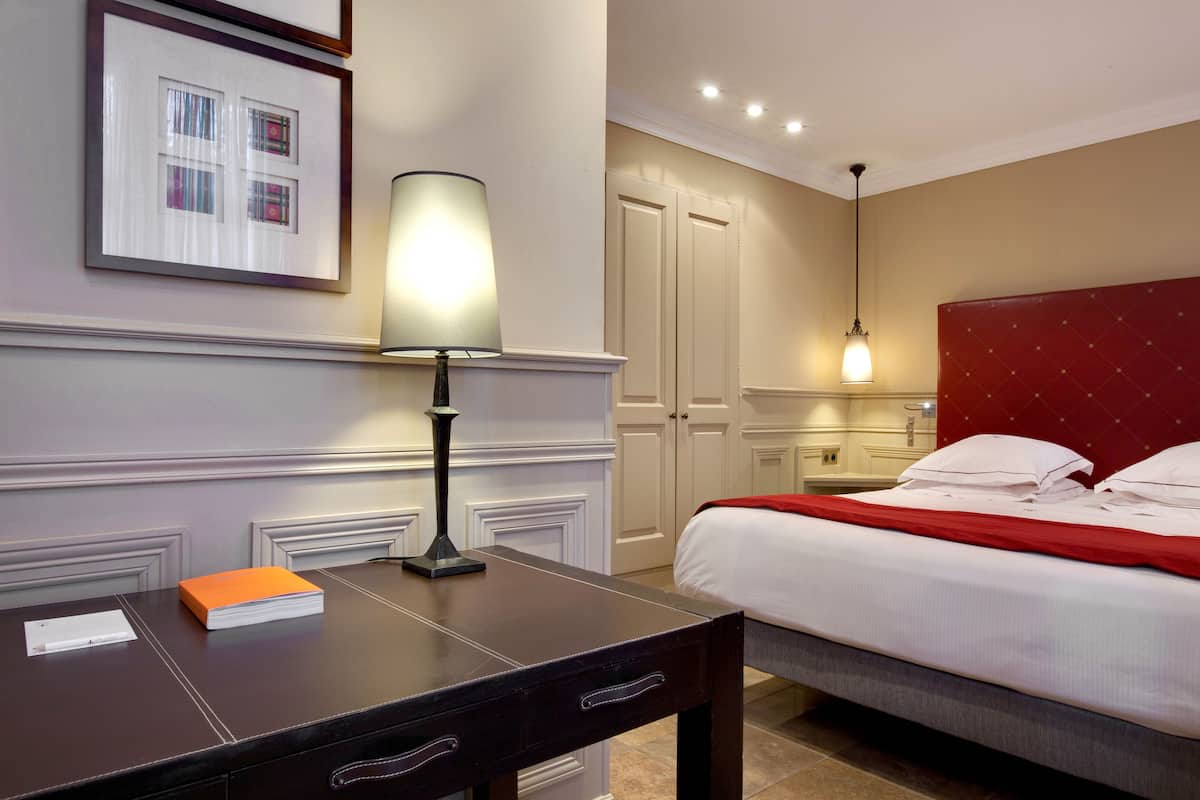 A hotel room with a neatly made bed, a bedside table with a lamp, a framed picture on the wall, and a door leading to a bathroom.