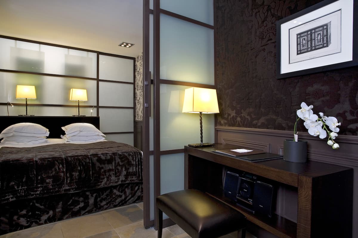 A hotel room with a bed, two bedside lamps, a desk with a computer, a small bench, and a framed picture on the wall.