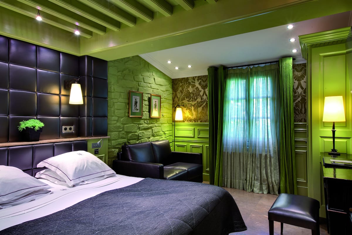 A hotel room with a bed, a seating area with a sofa and armchairs, a desk with a chair, and a window with curtains.