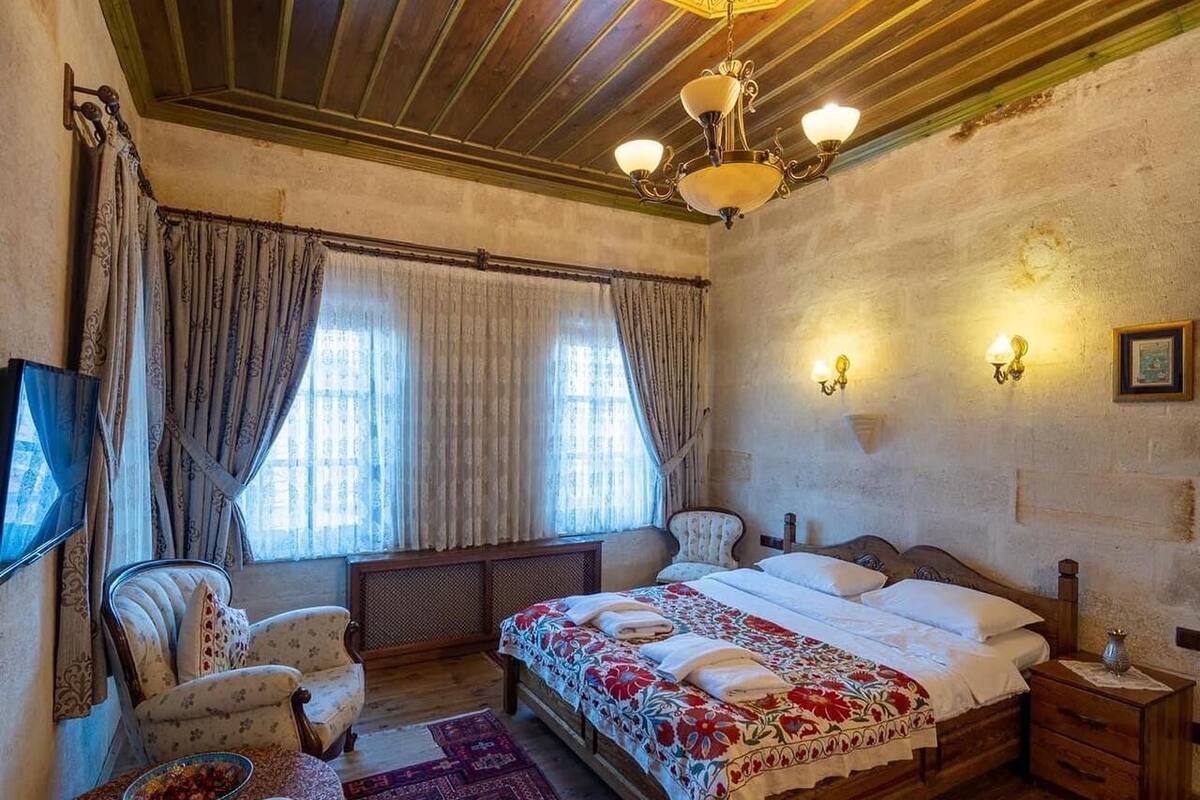 A traditional bedroom with a wooden ceiling, a large bed with floral bedding, two armchairs, a wooden nightstand, and a television on the left.