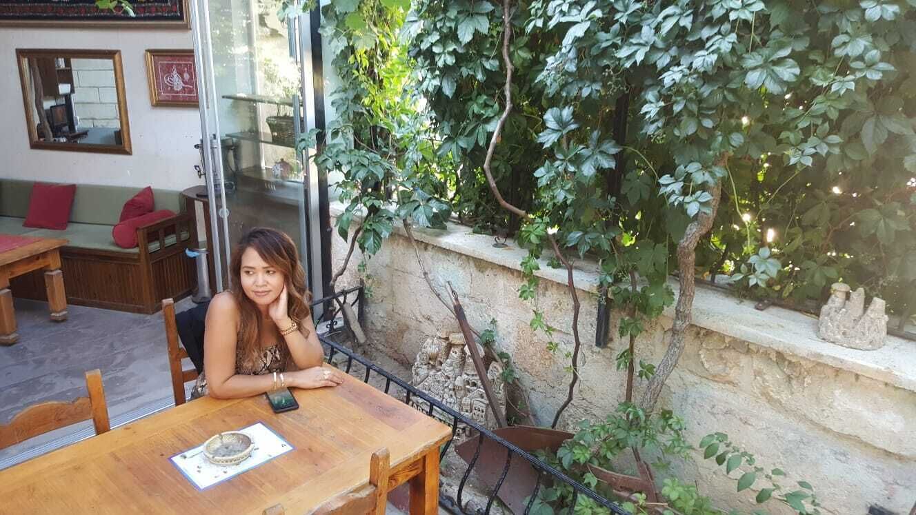 Una persona sentada a una mesa de madera al aire libre, con un muro de piedra y vegetación de fondo.