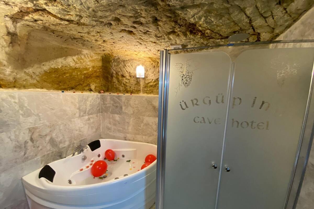A bathroom with a bathtub containing red balloons, a shower area, and a wall with the inscription "URGÜPO in cave hotel".