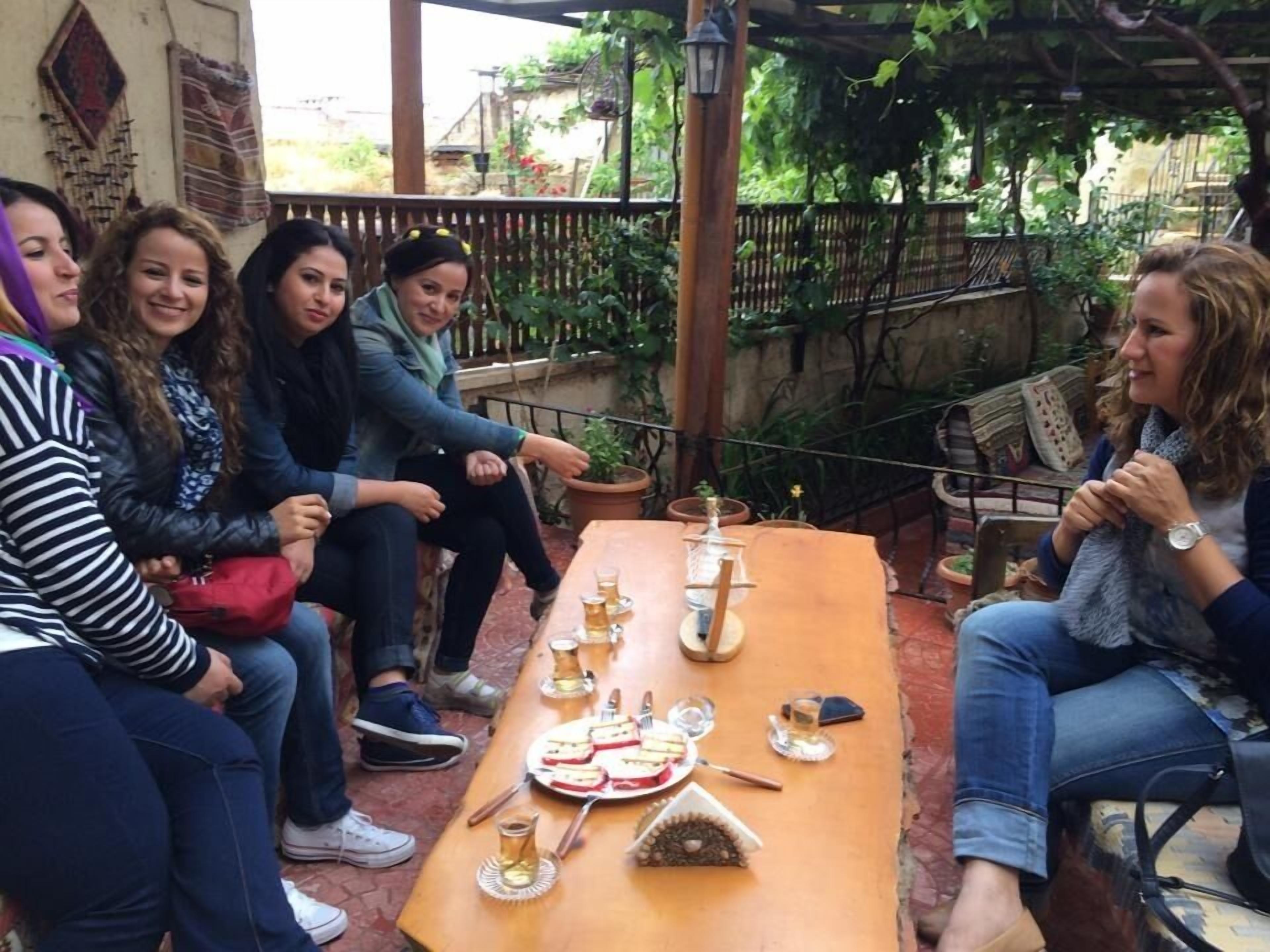 Un grup de persones assegudes al voltant d'un taula de fusta amb menjar i begudes, en un espai exterior amb una pèrgola de fusta i plantes en testos.