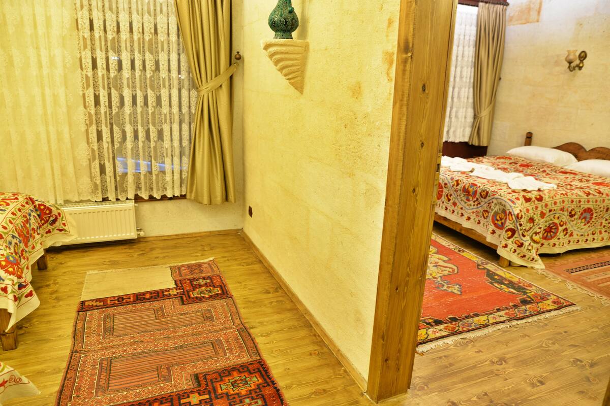 A room with a wooden floor, a bed with a floral bedspread, a red patterned rug, and a window with sheer curtains.