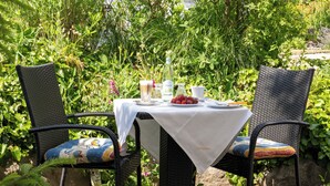 An outdoor dining area with wicker chairs and a table set with a white tablecloth, surrounded by lush greenery and plants. - Schöngarten Garni (Lindau)