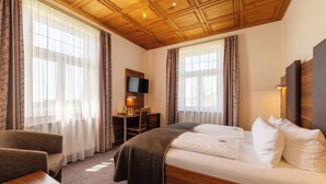 A hotel room with a wooden ceiling, a bed with white bedding, a desk with a chair, and a television mounted on the wall. - Schöngarten Garni (Lindau)