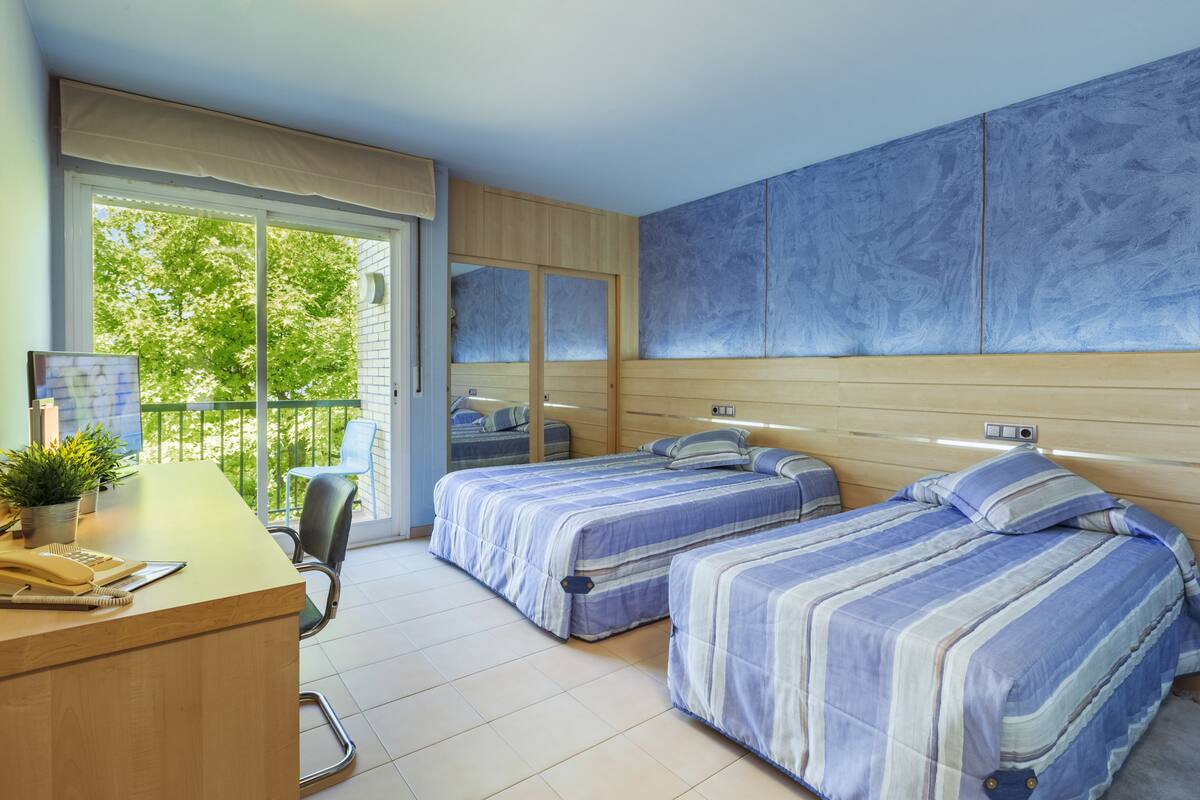 Ein Hotelzimmer mit zwei Betten, einem Schreibtisch, einem Stuhl und einem Balkon mit Blick auf Bäume.