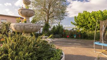 Surrounded by lush greenery, this serene outdoor space features a multi-tiered fountain and a playground with a slide and swing set.