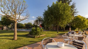 Un patio exterior con mesas y sillas de metal, rodeado de césped y árboles bien cuidados.