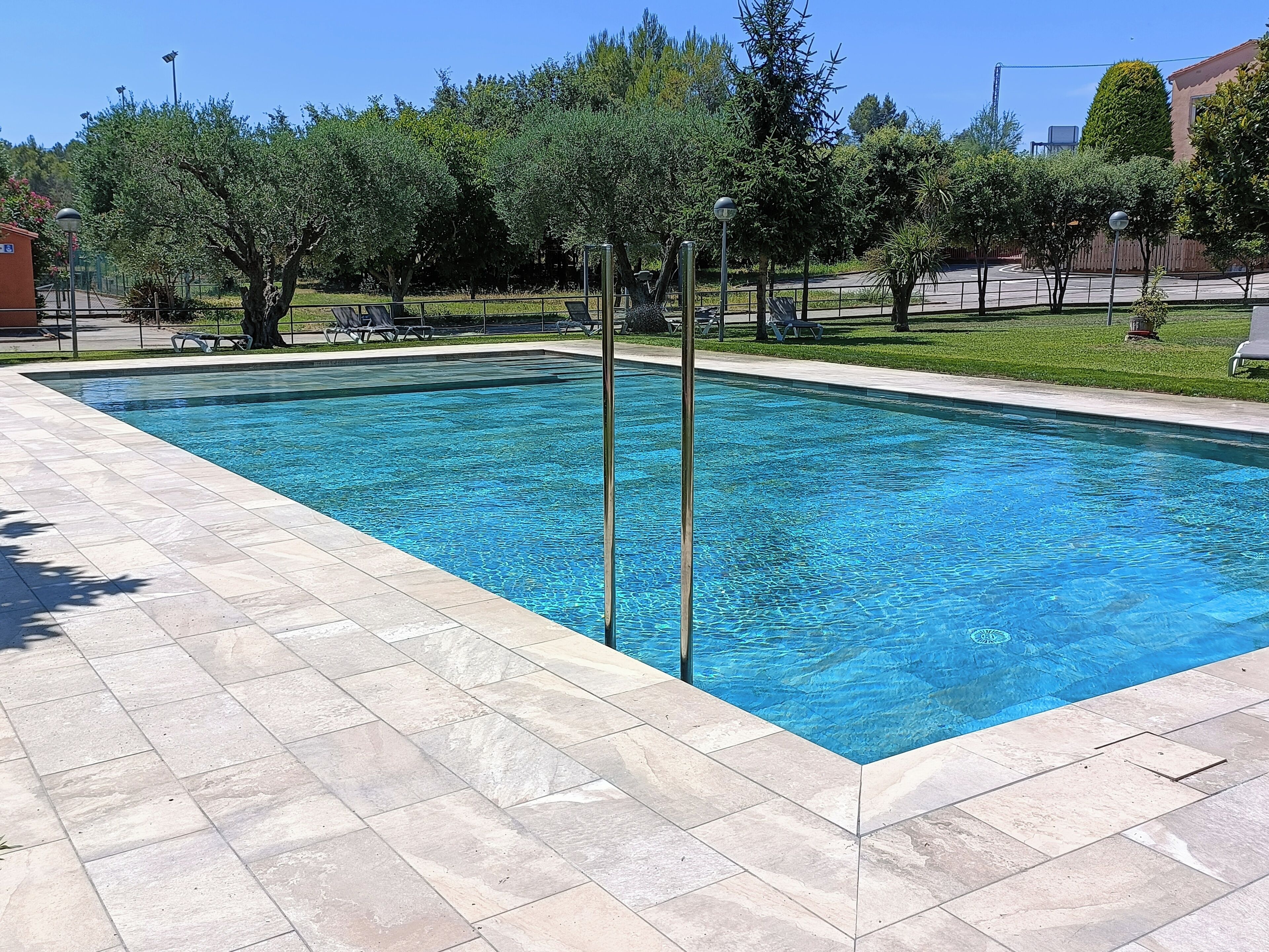 Une piscine rectangulaire avec une terrasse carrelée, entourée d’arbres et avec un bâtiment visible au loin.