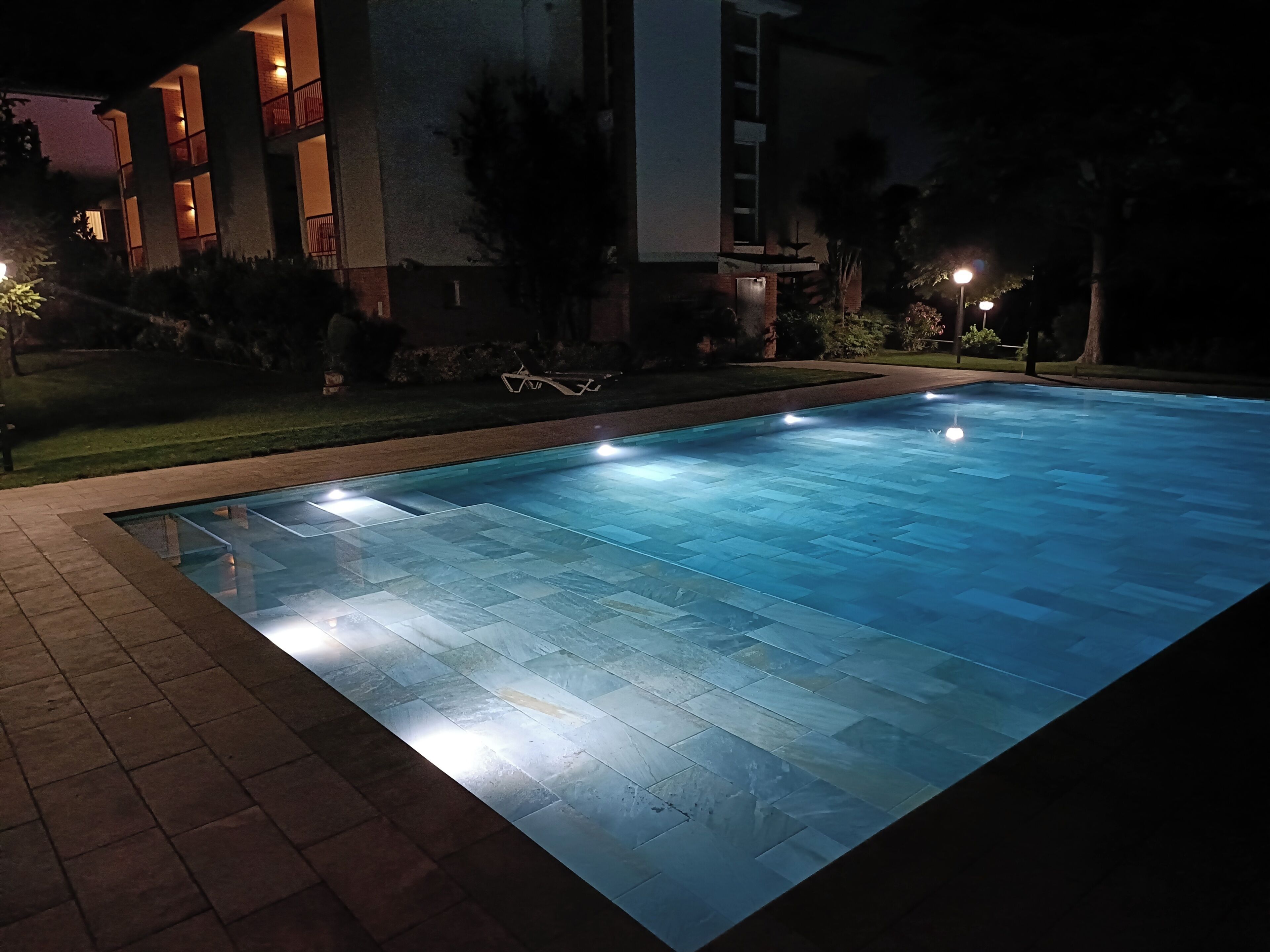 A swimming pool with a tiled edge, surrounded by a brick walkway, and adjacent to a building with multiple windows and balconies.