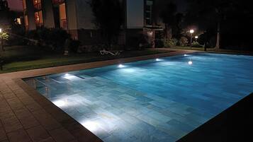 A swimming pool with a tiled edge, surrounded by a brick walkway, and adjacent to a building with multiple windows and balconies.