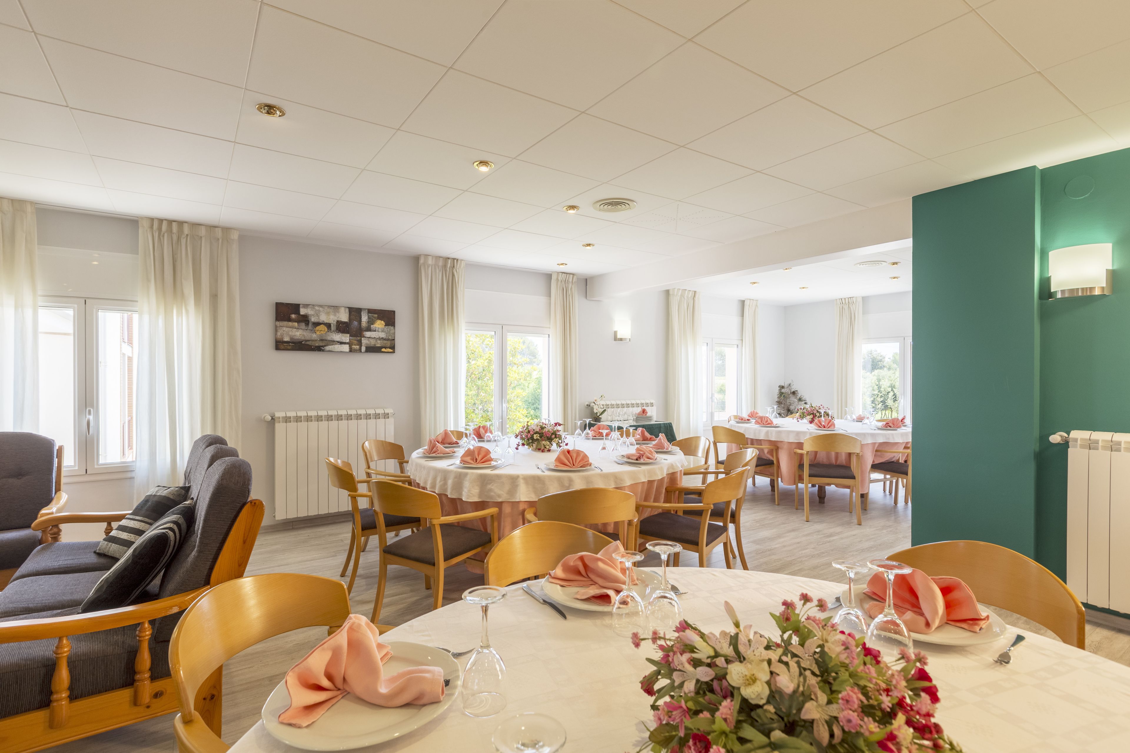 Un comedor con mesas redondas dispuestas para cuatro comensales, cada una con manteles blancos, servilletas rosas y centros de mesa florales. Rodeando las mesas, sillas de madera, y un sofá con cojines en primer plano.