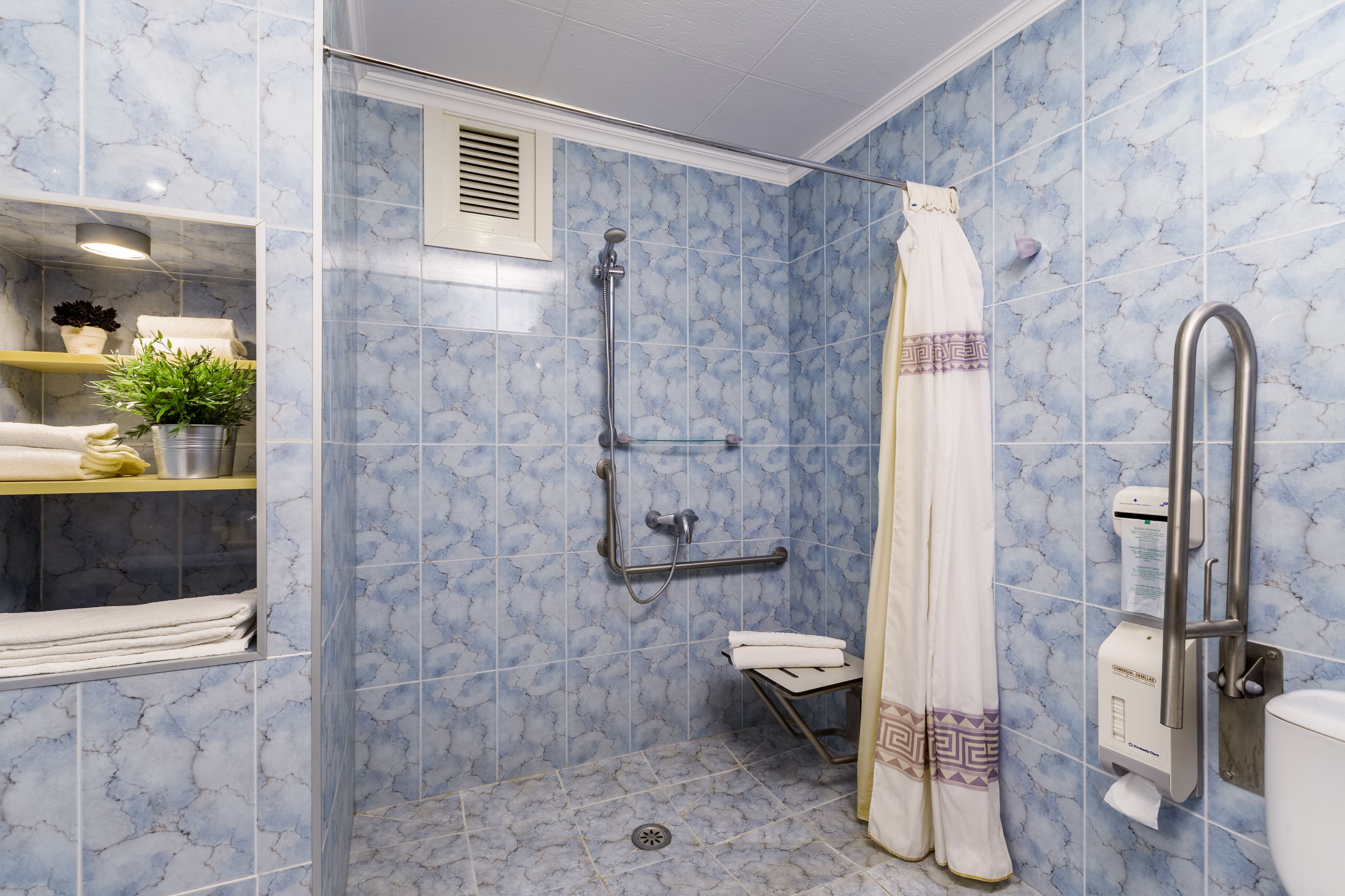 A bathroom with a shower, toilet, and a shelf with towels and plants.