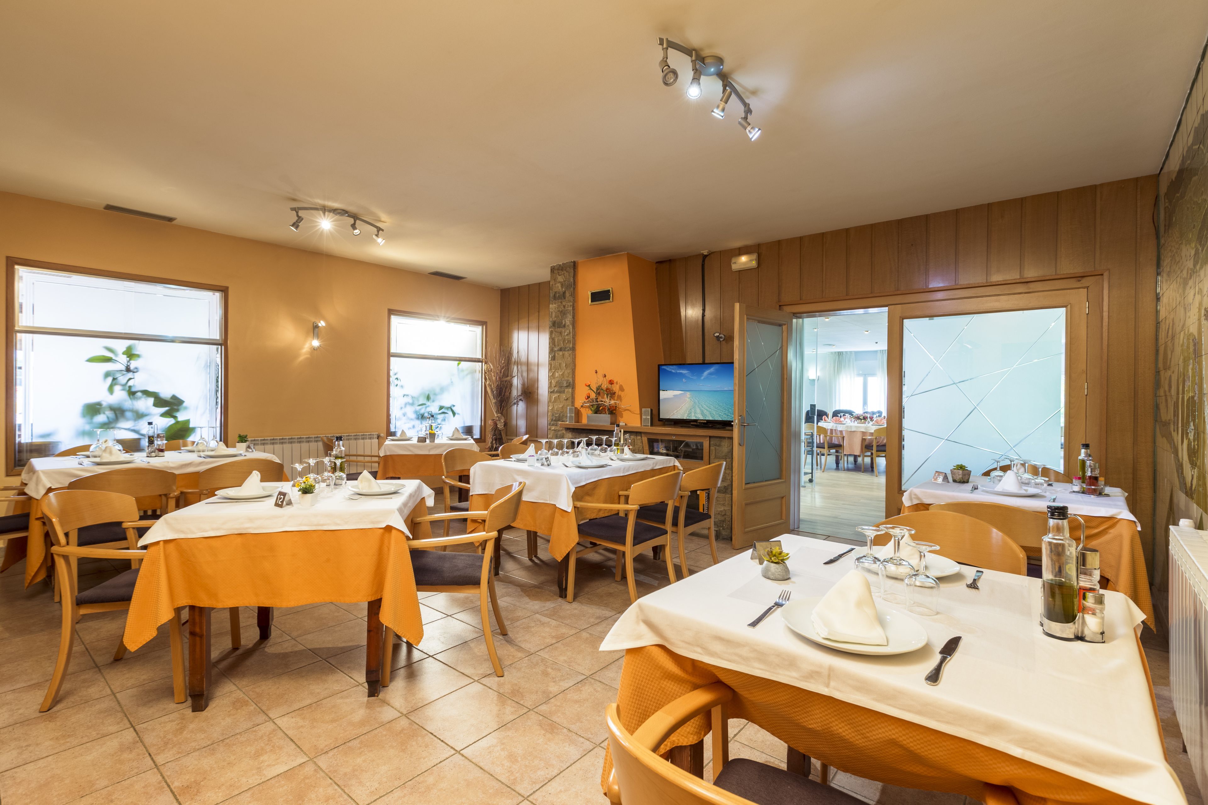 A dining area with multiple tables set for meals, a television on the wall, and large windows allowing natural light.