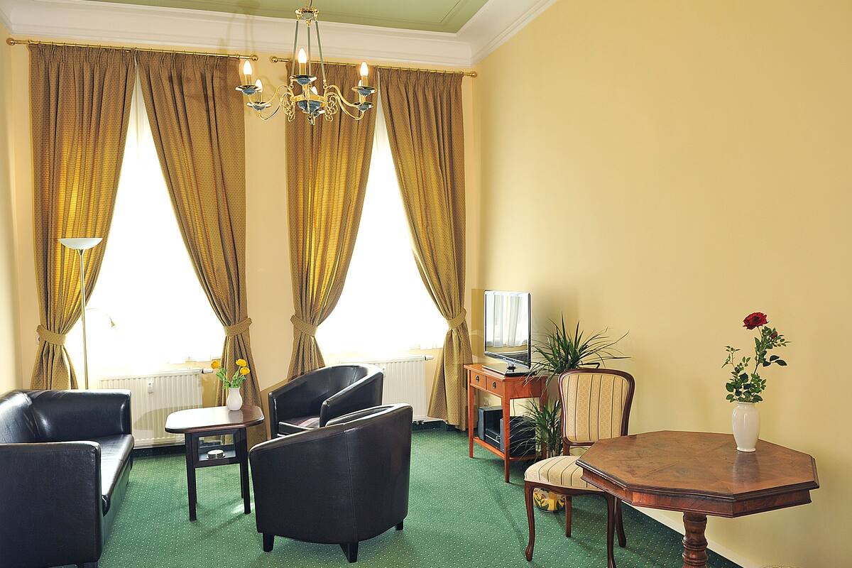 Hotelkamer met groen tapijt, twee zwarte leren fauteuils, een houten tafel, een klein bureau met een computer, een potplant en een vaas met een enkele rode roos.