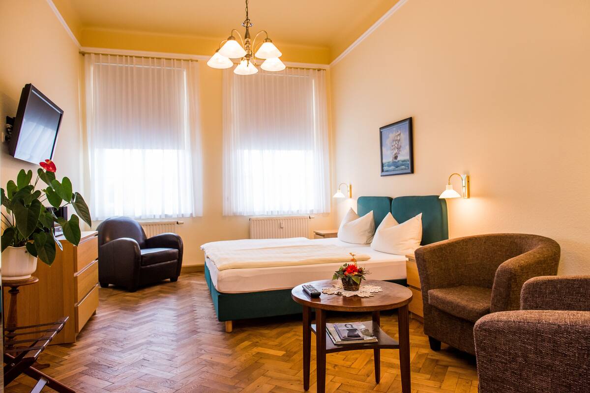 Hotelkamer met een bed, twee stoelen, een klein tafeltje, een kamerplant, een televisie en een raam met gordijnen.