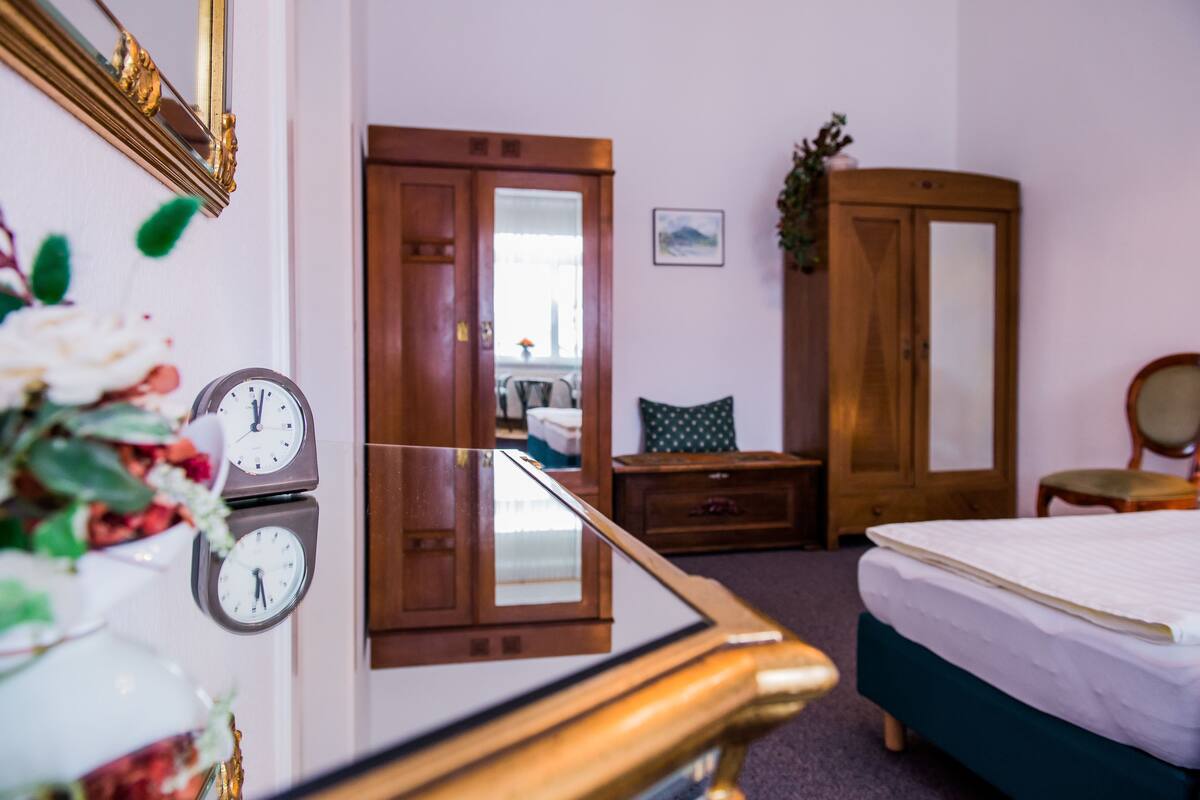 Een hotelkamer met een bed, een houten garderobe, een spiegel, een klok en een klein tafeltje met een bloevas.