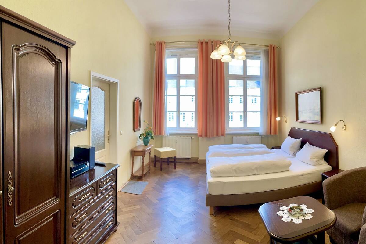 Een hotelkamer met een houten dressoir, een bed met witte lakens, een stoel, een klein tafeltje met een kantje en een raam met gordijnen.