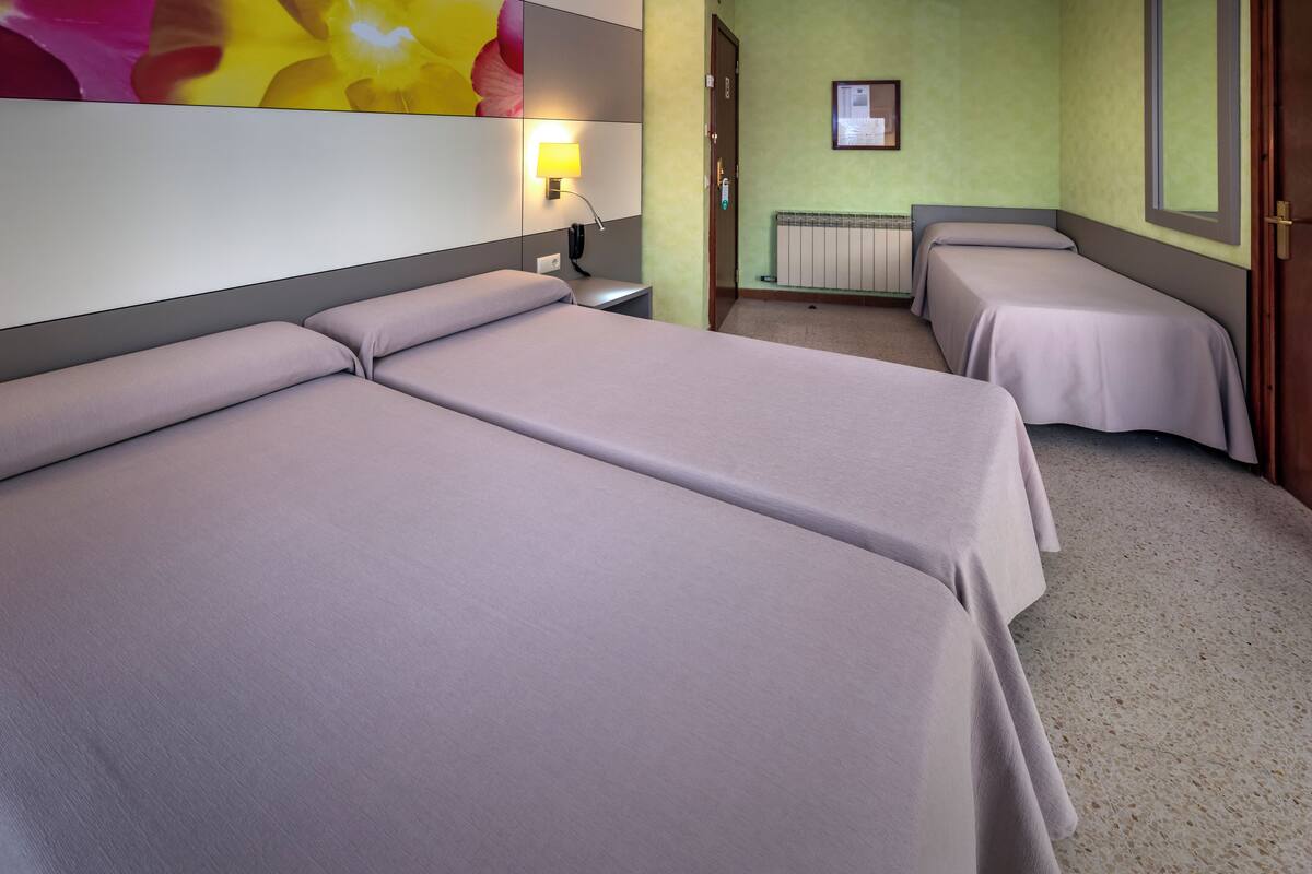 Habitación de hotel con dos camas, una mesita de noche, una lámpara, un cuadro enmarcado en la pared y una ventana.