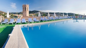 A pool with blue and white striped lounge chairs, a tower, and a view of a hillside town. - Hotel Sabiote (Pineda de Mar)