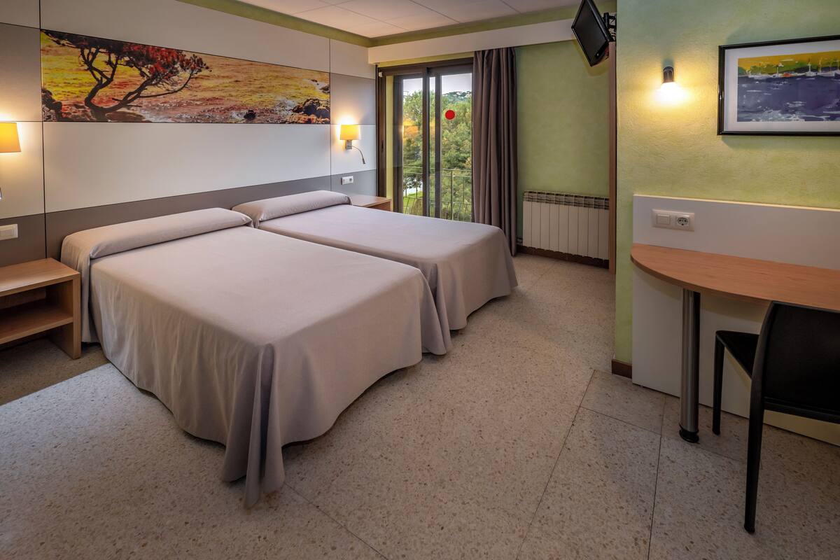 Habitación de hotel con dos camas, un escritorio, una silla, un televisor y una ventana con vista a árboles.