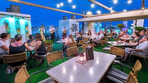 An outdoor dining area with wicker chairs and tables, a stage with a guitarist and singer, and a city view in the background. - Hotel Sabiote (Pineda de Mar)