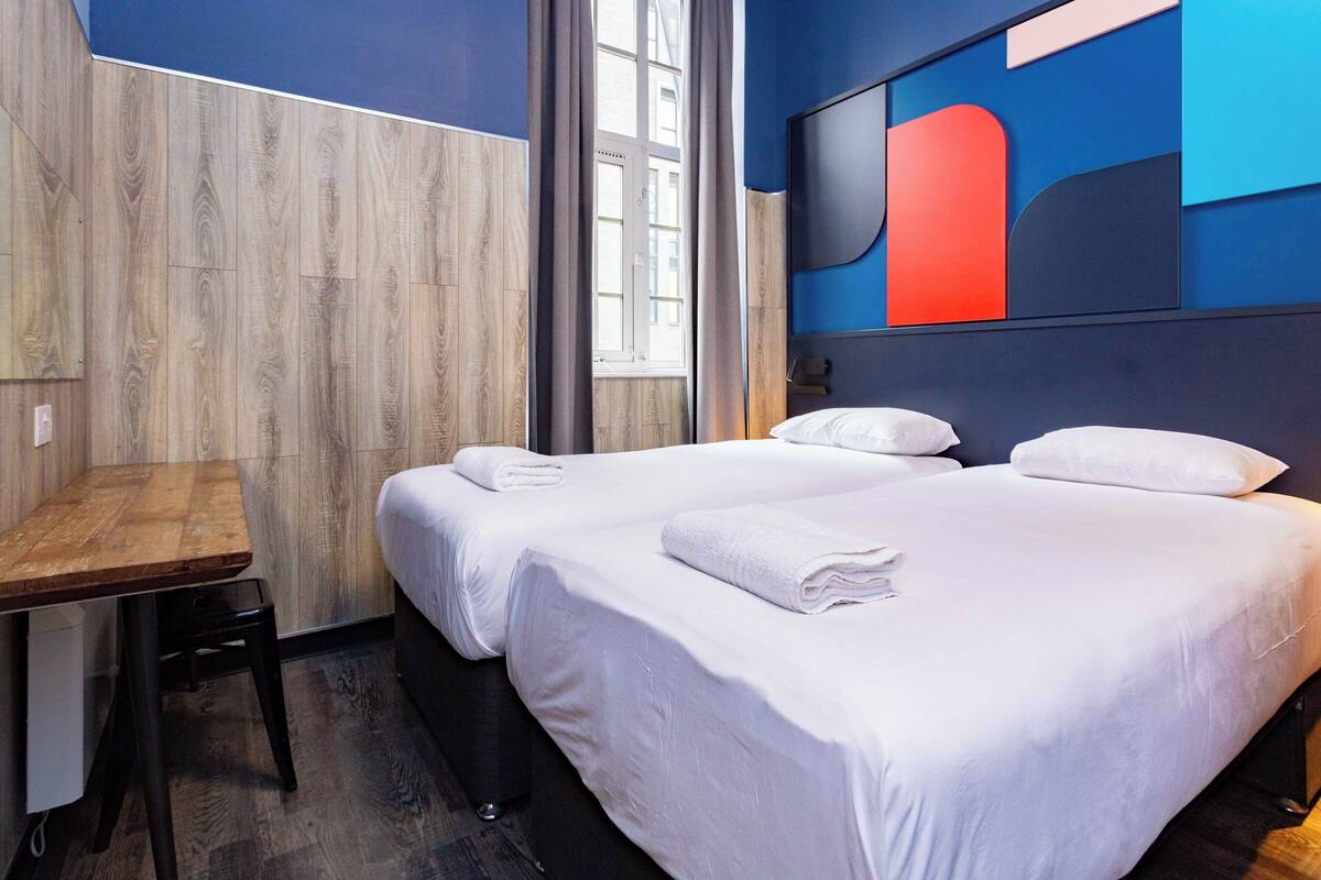 A hotel room with two beds, a wooden desk, and a large window.