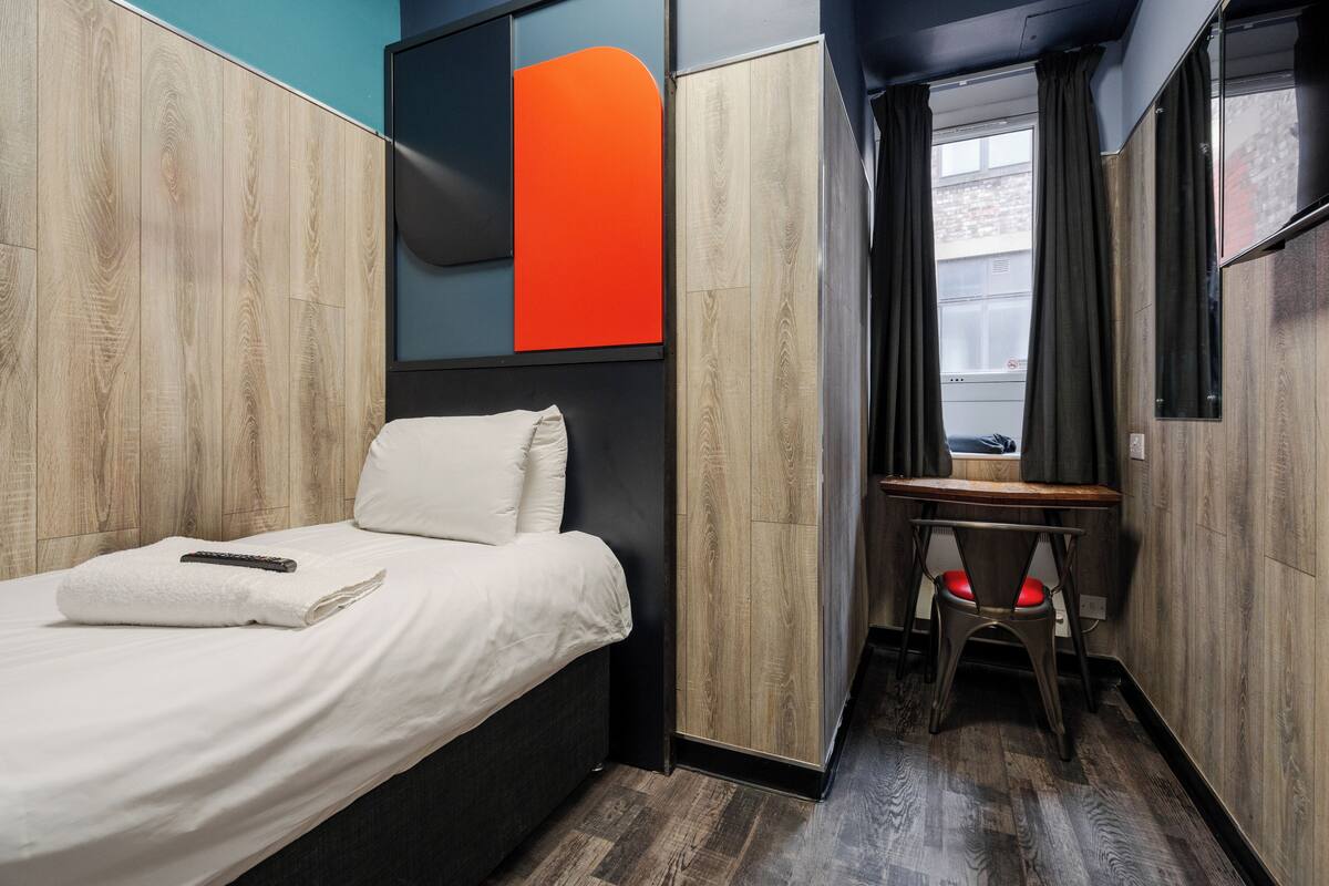 A compact hotel room with a bed, a desk, and a view of the city through a window.