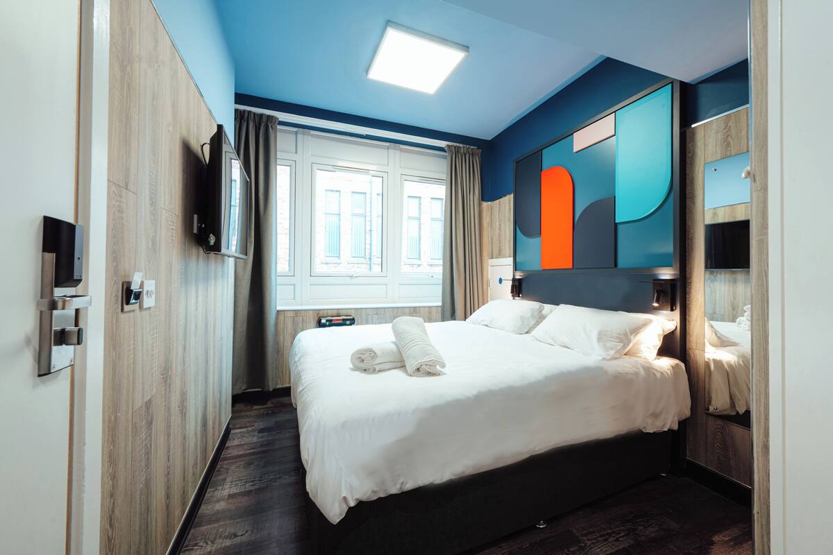 A modern hotel room with a large bed, a flat-screen TV, a window with curtains, and a unique wall art piece.