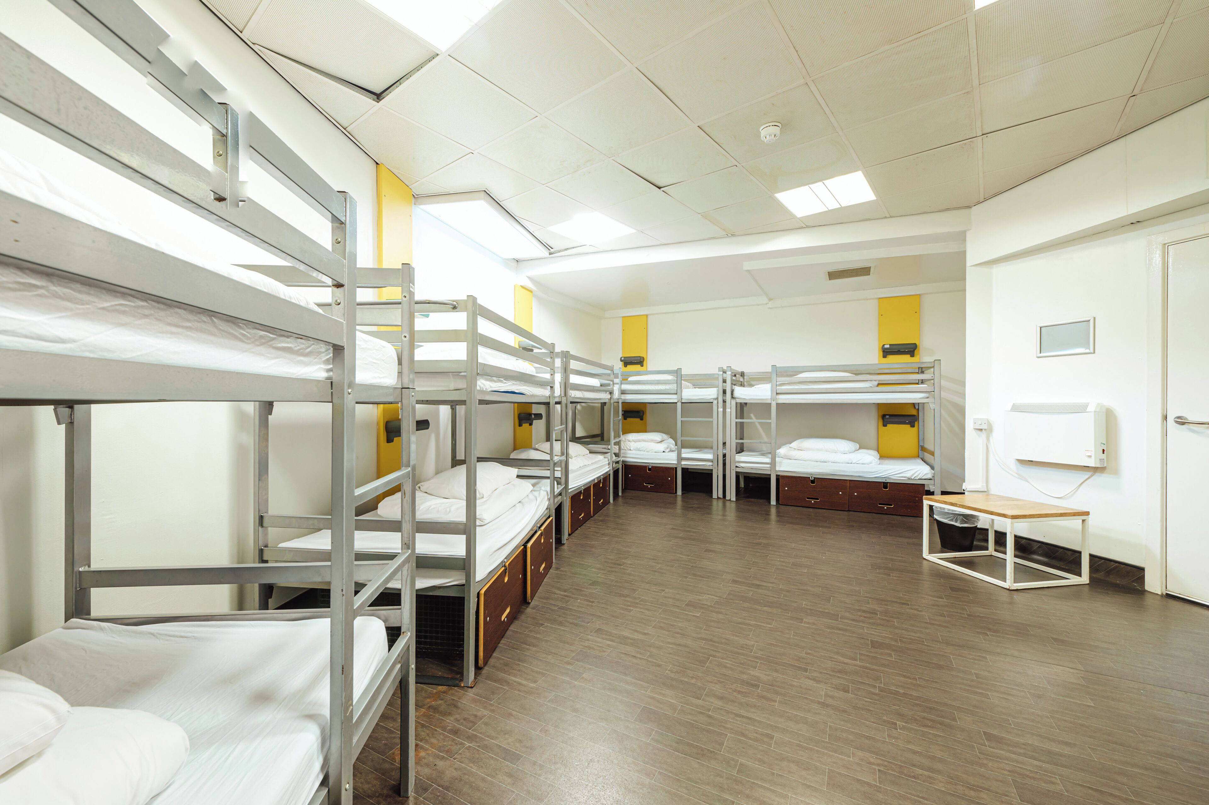 A dormitory room with bunk beds, a wooden table, and a trash bin.