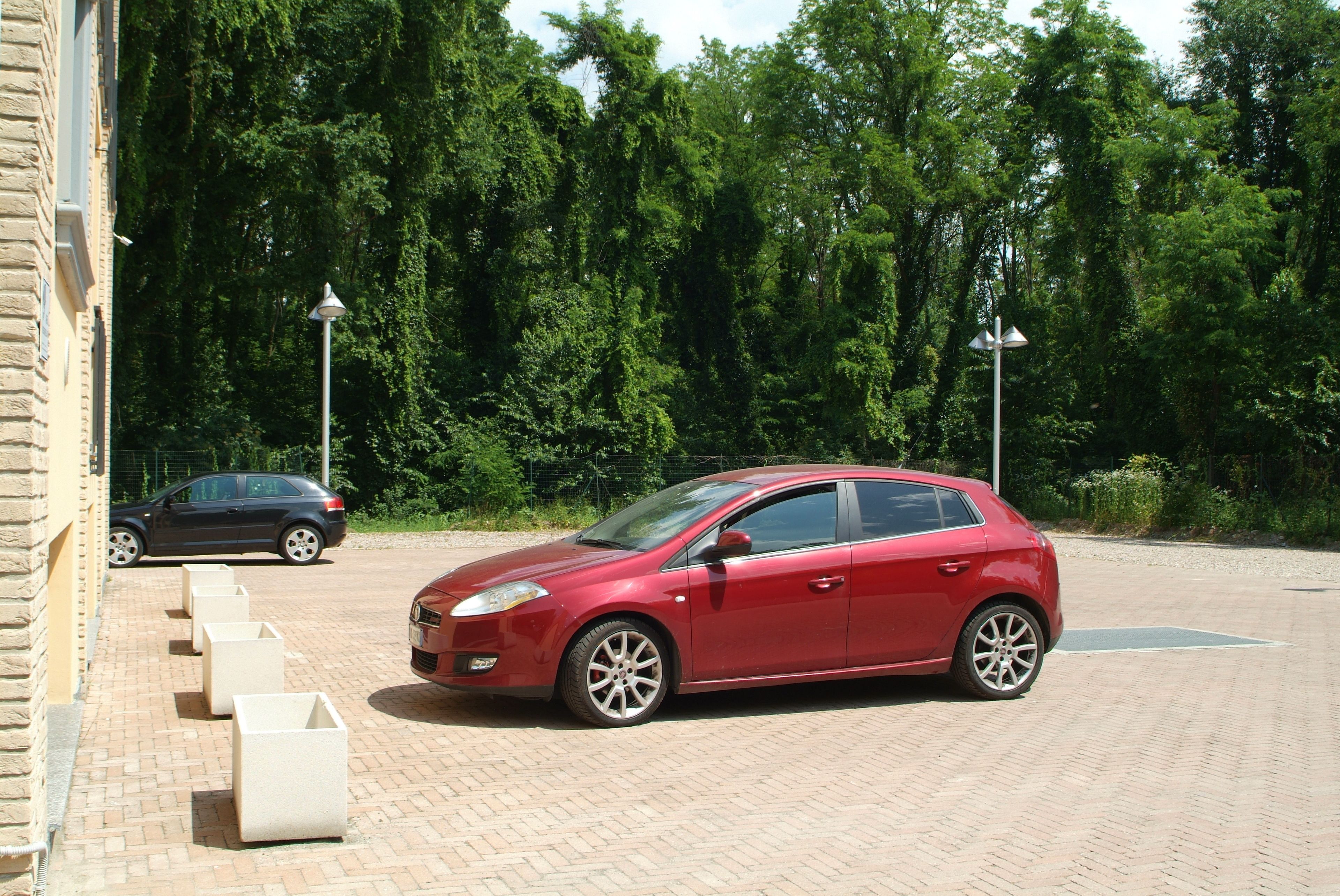 A red car parked on a paved area with a brick wall and trees in the background.