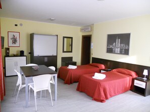 A hotel room with two beds, a dining table with chairs, a small kitchenette, and a cityscape picture on the wall. - San Prospero (Garbagnate Milanese)
