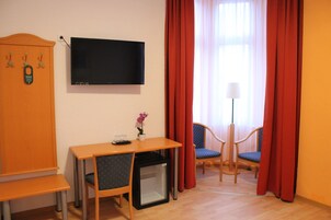 Habitación de hotel con televisión montada en la pared, una mesita con una silla, un refrigerador pequeño y dos sillones con cortinas rojas.