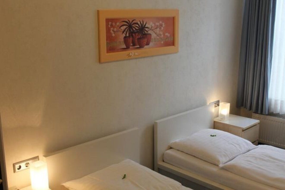Une chambre d’hôtel avec deux lits simples, une table de chevet avec une lampe, un tableau encadré au mur et une fenêtre avec des rideaux.