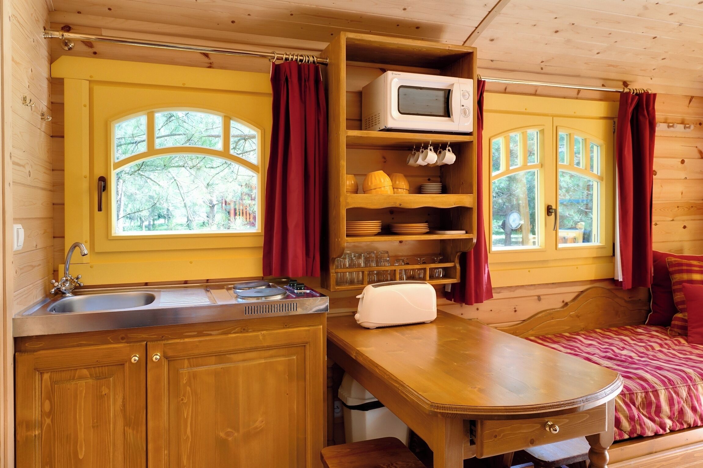 Interior de una cabaña de madera con cocina, una mesa de comedor y una cama con cortinas rojas.