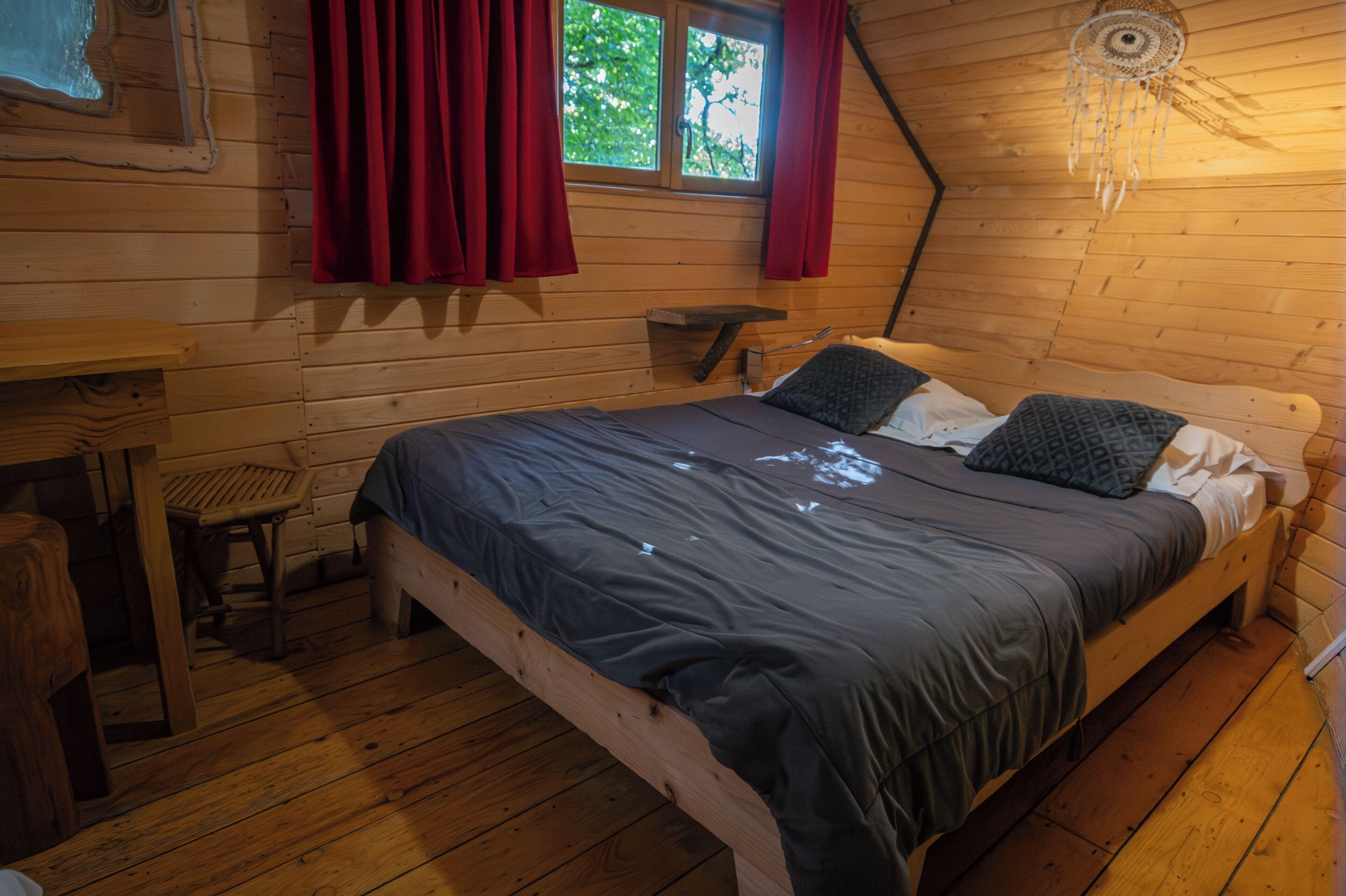A wooden cabin room with a bed, a small table, and a window with curtains.