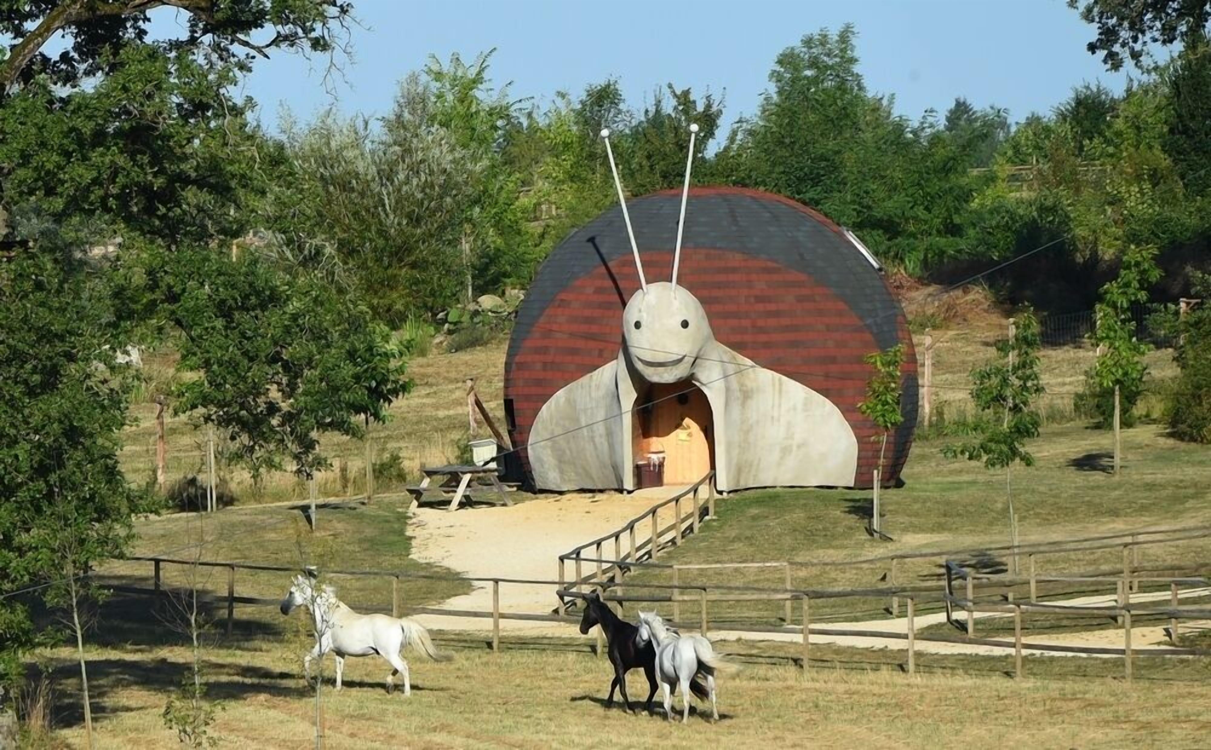 A large, circular structure resembling a giant insect with a tunnel entrance, surrounded by a fenced area with horses grazing.