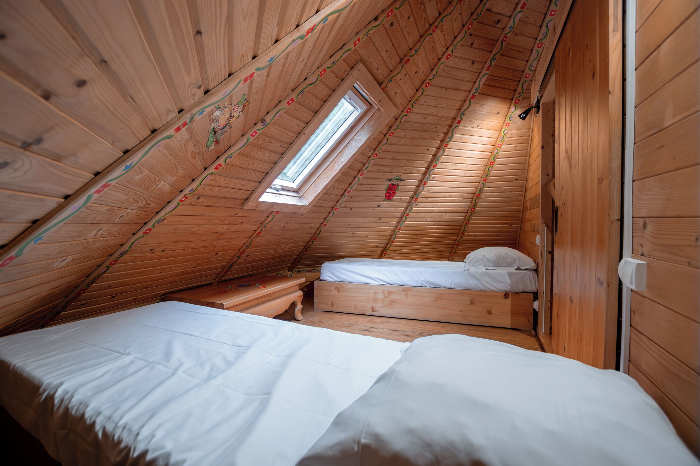 A small, wooden attic bedroom with two beds, a window, and a sloped ceiling.