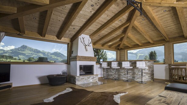 Un interior rústico con chimenea de piedra, vigas de madera, alfombra de cuero y vistas a la montaña.