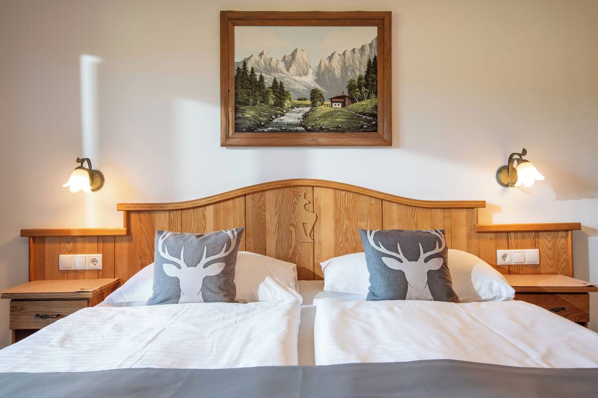 Ein Doppelbett mit weißer Bettwäsche und zwei Zierkissen, ein hölzernes Kopfende, Nachttische mit Wandlampen sowie ein gerahmtes Landschaftsbild über dem Bett.