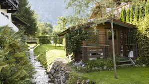 Una cabaña rústica con un camino de piedra que conduce a ella, rodeada de exuberante vegetación y un telón de fondo de montañas.