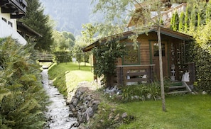 Un chalet rustique, accessible par un sentier de pierres, entouré d’une végétation luxuriante et adossé à une montagne.