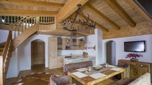 Un salón espacioso con una escalera de madera, una mesa de comedor con sillas acolchadas, una araña y una cocina al fondo.